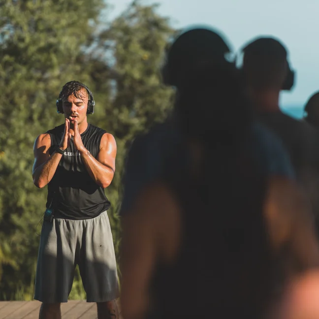 An instructor leads a mindful outdoor fitness session, hands together in front of his face. Participants join in, slightly out of focus in the foreground.