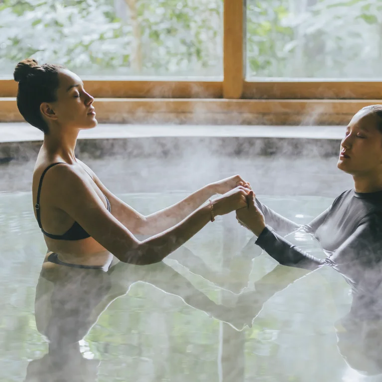 Khách hàng thư giãn trong một suối nước nóng trong nhà, tay trong tay và đối diện nhau. Những ô cửa sổ lớn khung cảnh cây cối xanh tươi xung quanh, tạo nên không gian yên bình và thư thái.