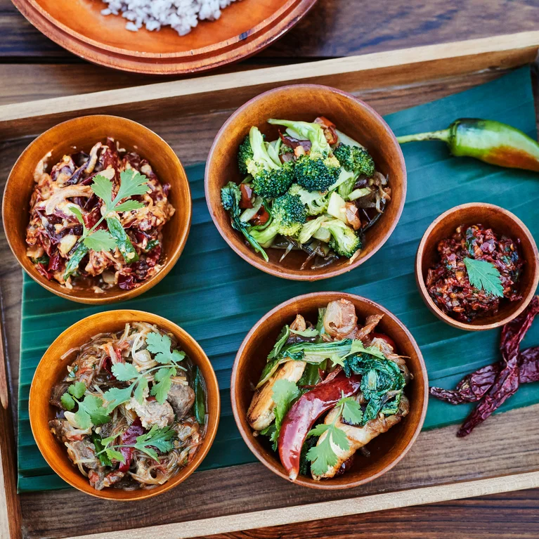 Cinq bols de cuisine thaïlandaise vibrante, chacun garni d'herbes fraîches, sont présentés sur un plateau tapissé de feuilles de bananier, accompagnés de piments rouges et verts.