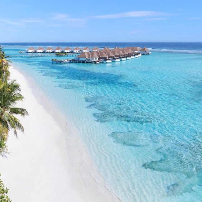 Overwater villas built with natural materials extend above calm blue water, bordered by sand and greenery under a clear sky. Eco-forward design integrates with the seascape.