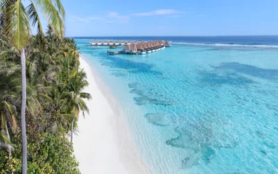 Overwater villas built with natural materials extend above calm blue water, bordered by sand and greenery under a clear sky. Eco-forward design integrates with the seascape.