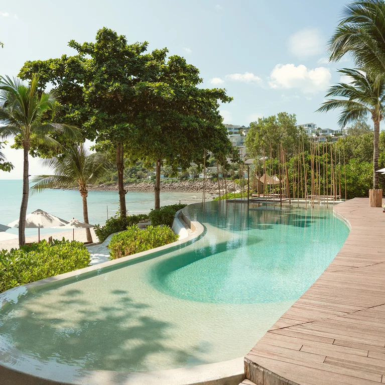 حوض سباحة بدرجات لطيفة، تظللها أشجار النخيل، ويحدها سطح خشبي وكراسي استرخاء تطل على المحيط والسماء المفتوحة. تضفي الألوان الطبيعية أجواءً هادئة.