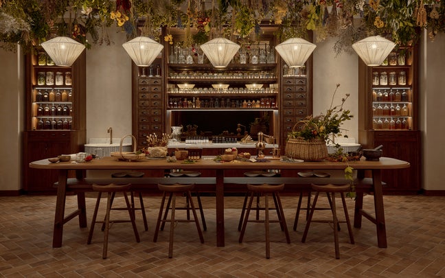 Spacious kitchen featuring a long wooden island, pendant lighting, stools, organized open shelving with jars, cabinets, and dried flowers hanging overhead.