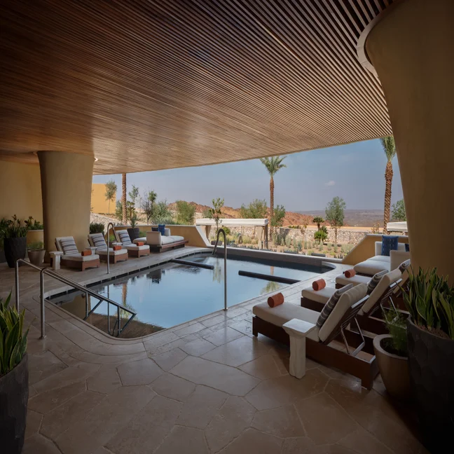 Indoor-outdoor pool area with lounge chairs, potted greenery, and warm wooden ceiling, open to palm trees and natural views for a relaxing and restorative atmosphere.