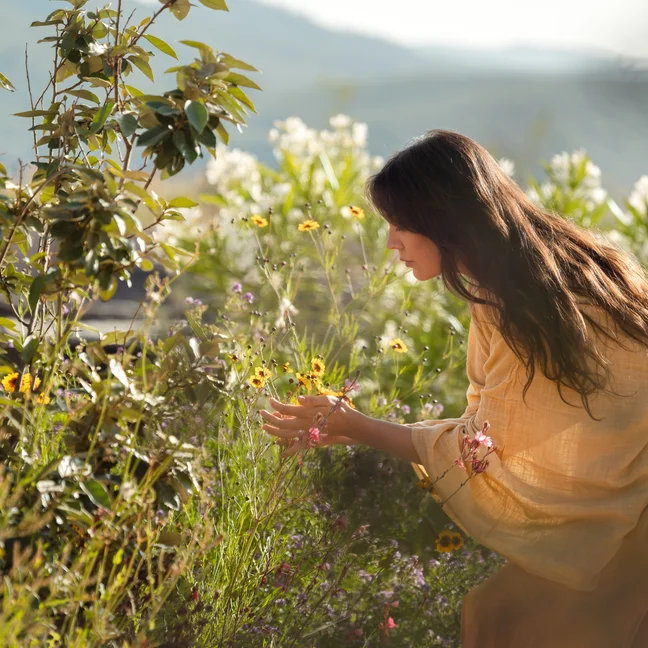 Guest in a yellow dress gathers wildflowers in a sunlit garden, among greenery and blooms, reflecting Six Senses’ connection to nature and mindful experiences.