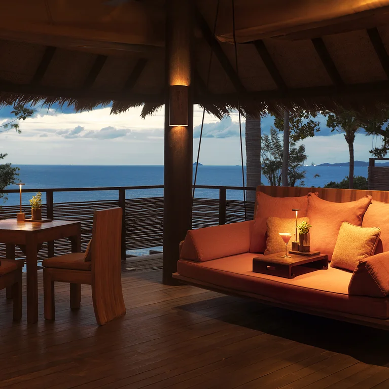 Le patio comprend une balançoire rembourrée et une table à manger en bois, avec vue sur l'océan au coucher du soleil et un éclairage doux et chaleureux, conçu pour le confort et la connexion avec la nature.