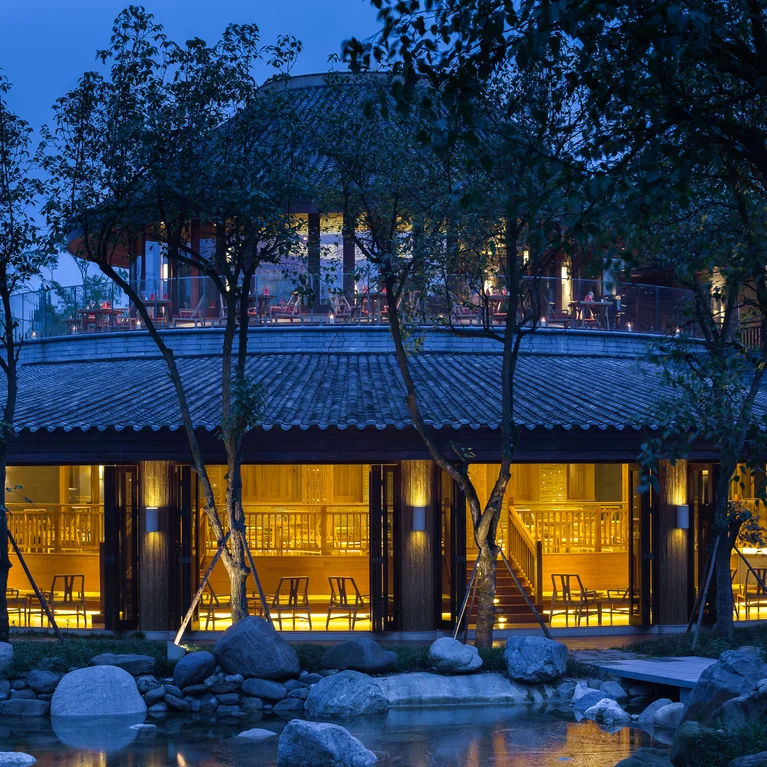 Un bâtiment circulaire avec de grandes fenêtres et des accents en bois se trouve à côté d'un étang tranquille, encadré par des arbres au crépuscule. Un éclairage doux crée une atmosphère calme et accueillante.