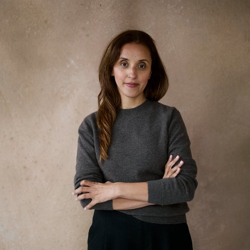 Person with long brown hair, wearing a gray sweater and black pants, stands before a softly textured neutral backdrop with arms comfortably crossed.