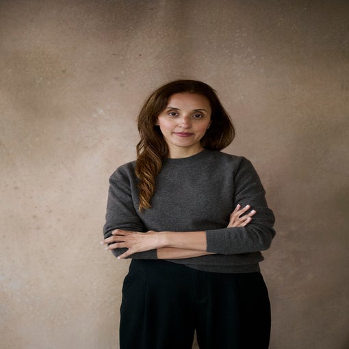 Person with long brown hair, wearing a gray sweater and black pants, stands before a softly textured neutral backdrop with arms comfortably crossed.