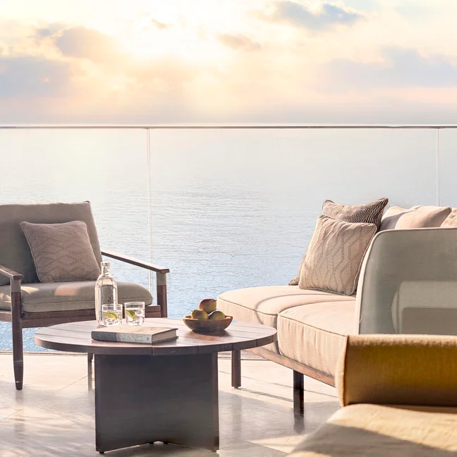 Balcony features natural seating, a table set with fruit and water, overlooking tranquil ocean views in evening light, designed for comfort and mindful relaxation.