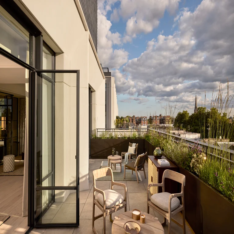 Private balcony featuring thoughtfully arranged outdoor seating, greenery in planters, and a cityscape view beneath a partly cloudy sky.