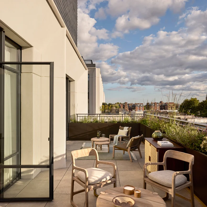 Private balcony featuring thoughtfully arranged outdoor seating, greenery in planters, and a cityscape view beneath a partly cloudy sky.