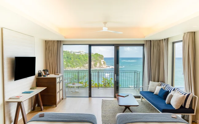 Ocean-facing room featuring beds, sofa, TV, and balcony. Floor-to-ceiling windows provide expansive views of cliffs and sea with abundant natural light.