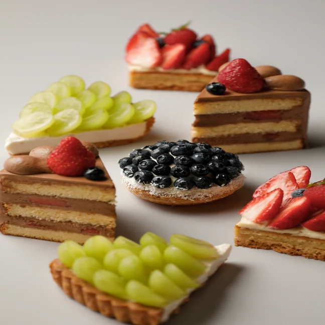 Fruit tarts and cakes topped with strawberries, blueberries, and grapes are neatly displayed in a circular pattern on a pale background.