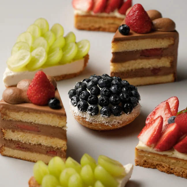 Fruit tarts and cakes topped with strawberries, blueberries, and grapes are neatly displayed in a circular pattern on a pale background.