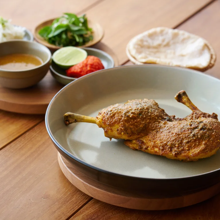 Roasted chicken served with sauce, fresh greens, a wedge of lime, sliced tomato, and artisanal flatbread on a wooden table setting.
