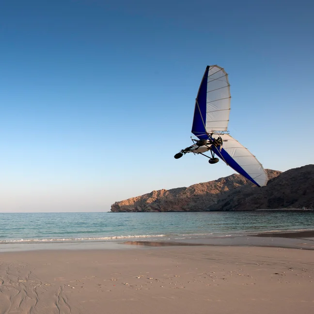 An ultralight aircraft glides above a sandy beach bordered by gentle waves and rocky cliffs, all beneath a clear sky.