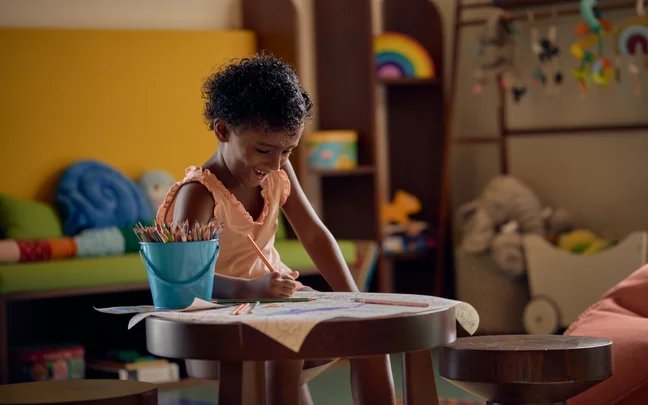 A child draws with colored pencils at a round table in a bright, welcoming room with toys and rainbows, designed to inspire creativity and comfort.