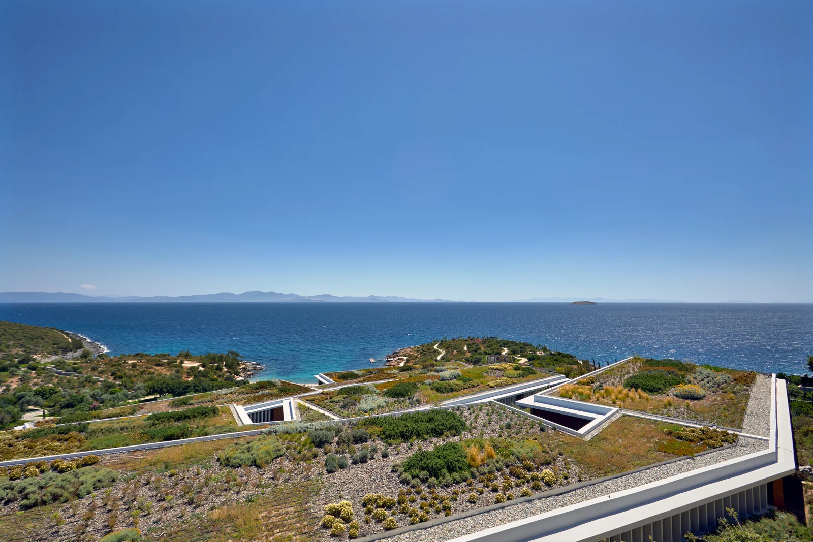 The resort's planted roofs seen from above, low white parapets and light wells set into rough scrub on a headland, with the Aegean and distant islands behind.