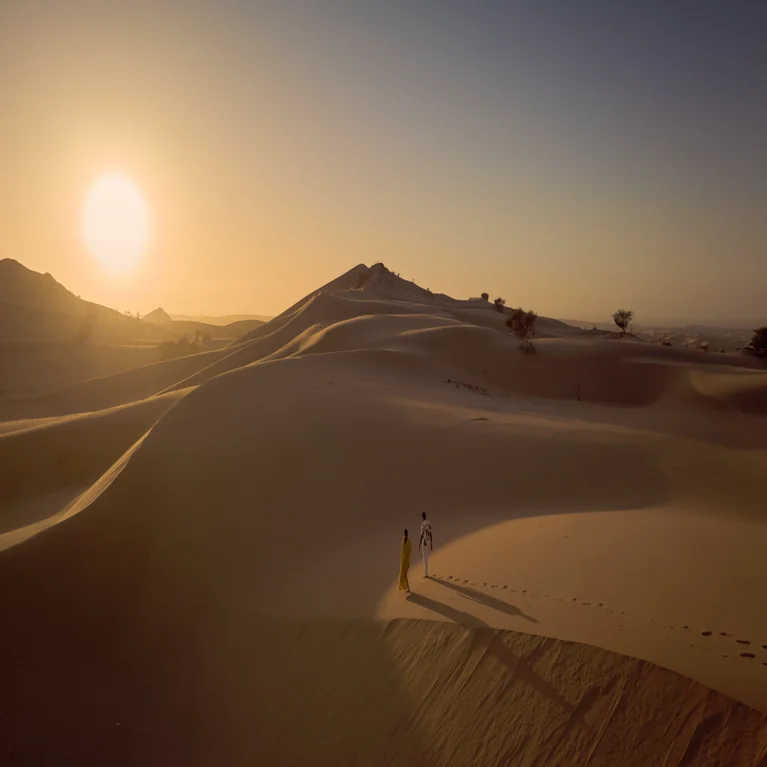 Guests journey across sand dunes in evening light, with clear sky above and sunlight creating gentle shadows, reflecting harmony with the natural desert environment.