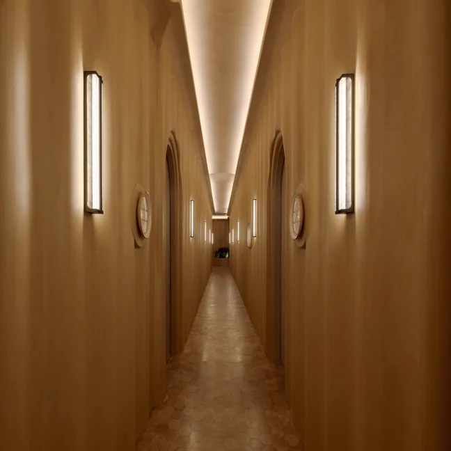 Beige-walled corridor with arched doorways and contemporary wall lighting. Soft illumination along the ceiling and floor highlights a calming, accessible passageway.