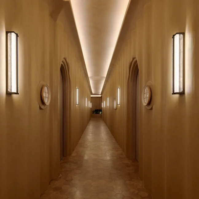Beige-walled corridor with arched doorways and contemporary wall lighting. Soft illumination along the ceiling and floor highlights a calming, accessible passageway.