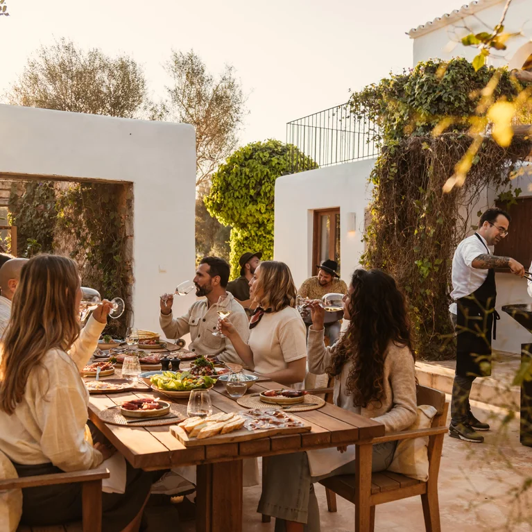 白天,在绿树和白色建筑的掩映下,客人在木桌上露天用餐,两位厨师在旁边的烤架上准备食物。.