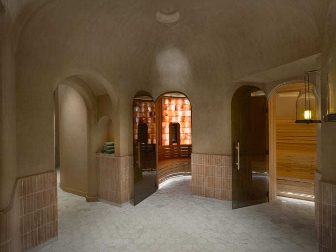 Calm interior features arched entryways, neutral walls, neatly arranged towels, and a sauna with salt block walls, seen through clear glass doors.