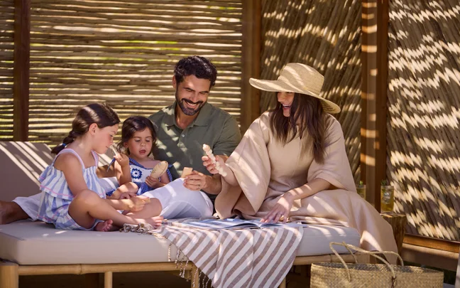 A family sits on a daybed in a shaded wooden structure, playing with wooden blocks and looking at a book together.