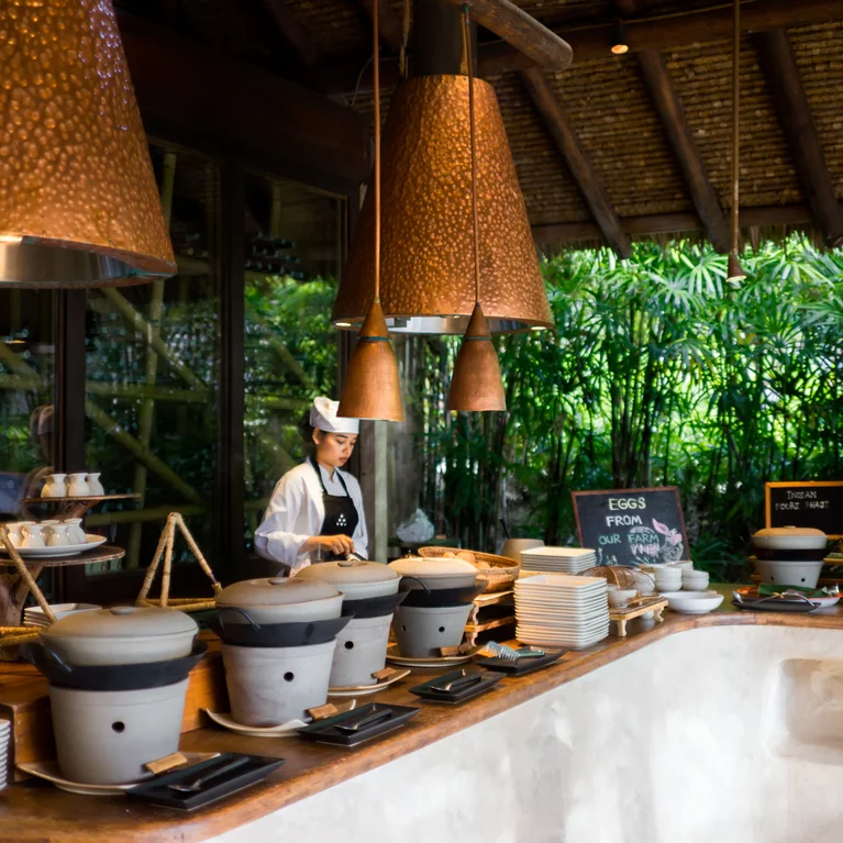 Le buffet du petit-déjeuner propose des plats artisanaux dans des pots et des assiettes, clairement étiquetés, sous de chaudes lampes en cuivre. De grandes fenêtres offrent une vue sur la verdure luxuriante.