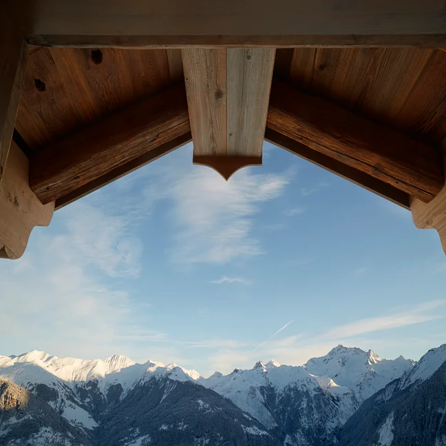 Pics montagneux enneigés et ciel dégagé à travers les poutres en bois du toit d'un chalet, vus sous l'avant-toit.
