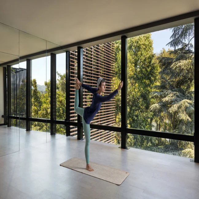 Guest doing yoga on a mat in a light-filled contemporary room with floor-to-ceiling windows and views of surrounding greenery.