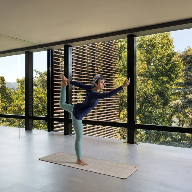 Guest doing yoga on a mat in a light-filled contemporary room with floor-to-ceiling windows and views of surrounding greenery.