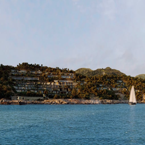 Eco-inspired villas set among green hills face a tranquil blue sea, with a sailboat passing by the shore under clear skies.