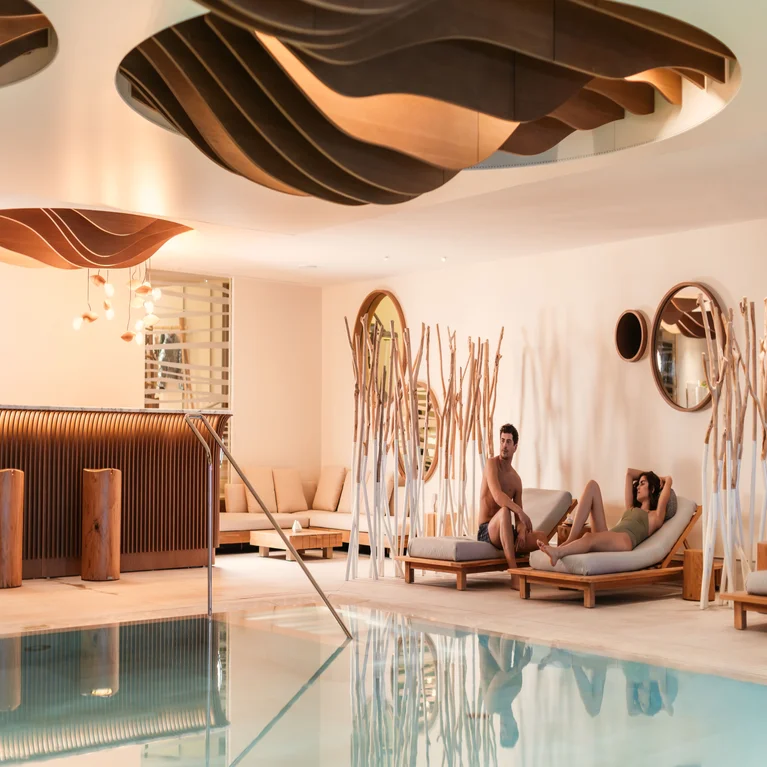 A man and a woman rest on lounge chairs by an indoor swimming pool, framed by screens made of wooden branches.