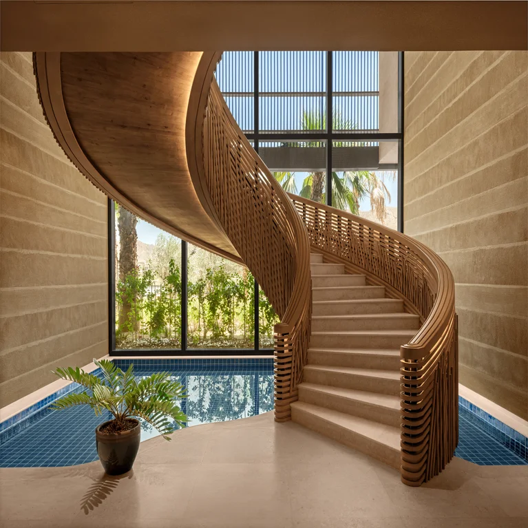 Wooden staircase rises above an indoor pool. A potted plant sits nearby, with large windows offering a view of palm trees outside.