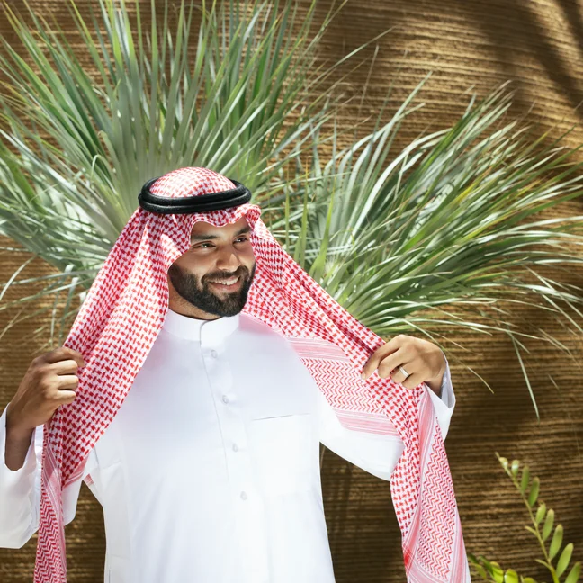 A man in traditional Middle Eastern dress stands by a palm tree, gently holding his red and white keffiyeh, sunlight creating soft patterns behind him.
