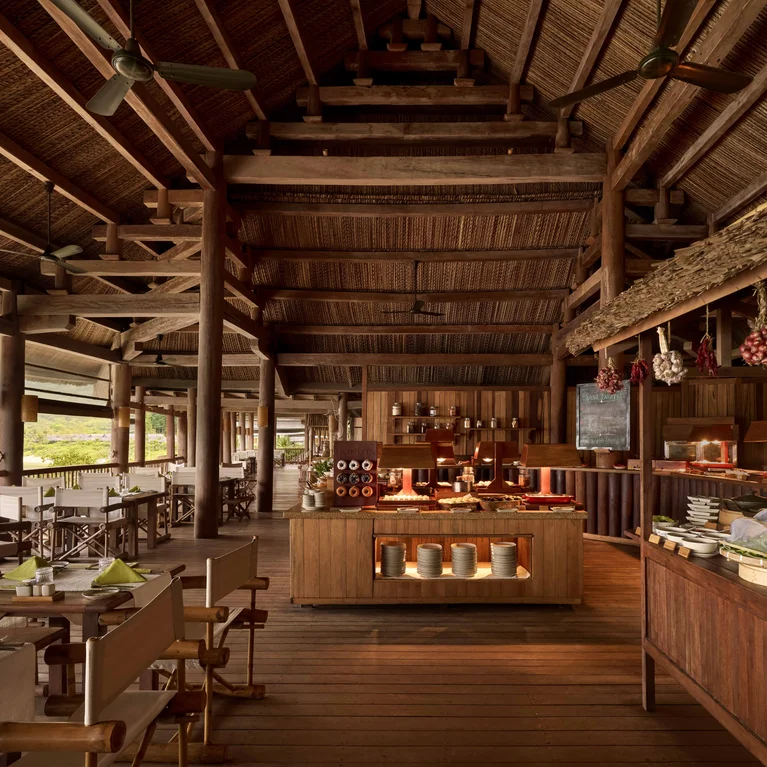 Open-air restaurant with natural wood elements, buffet stations, hanging lights and tables set for dining; views of the surrounding vegetation enhance the space.