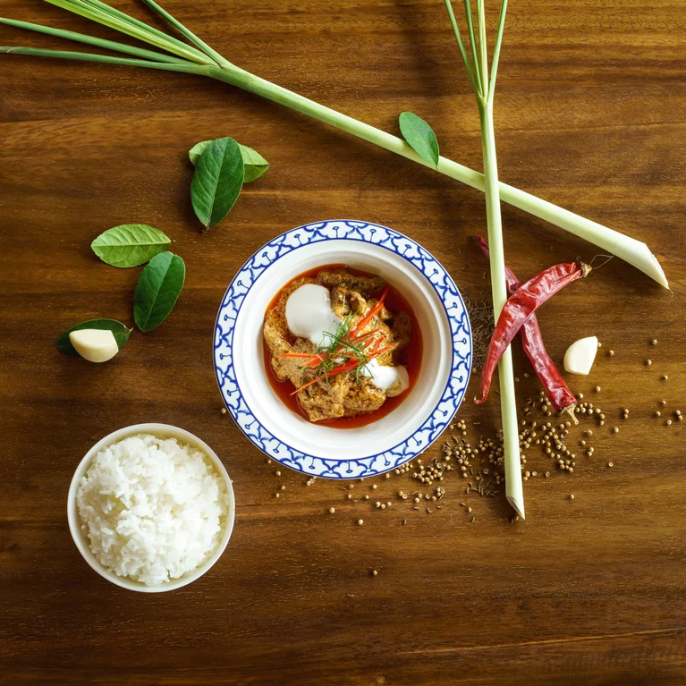 Thai curry served with white rice on a wooden table, accompanied by lemongrass, dried chilies, garlic, and kaffir lime leaves arranged alongside.