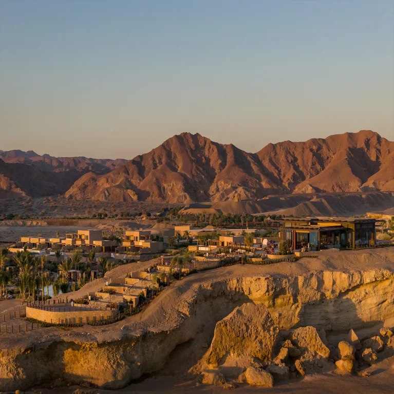 Sustainable architecture and palm trees stand at the foot of sunlit, rocky mountains in a serene desert landscape below an open sky.