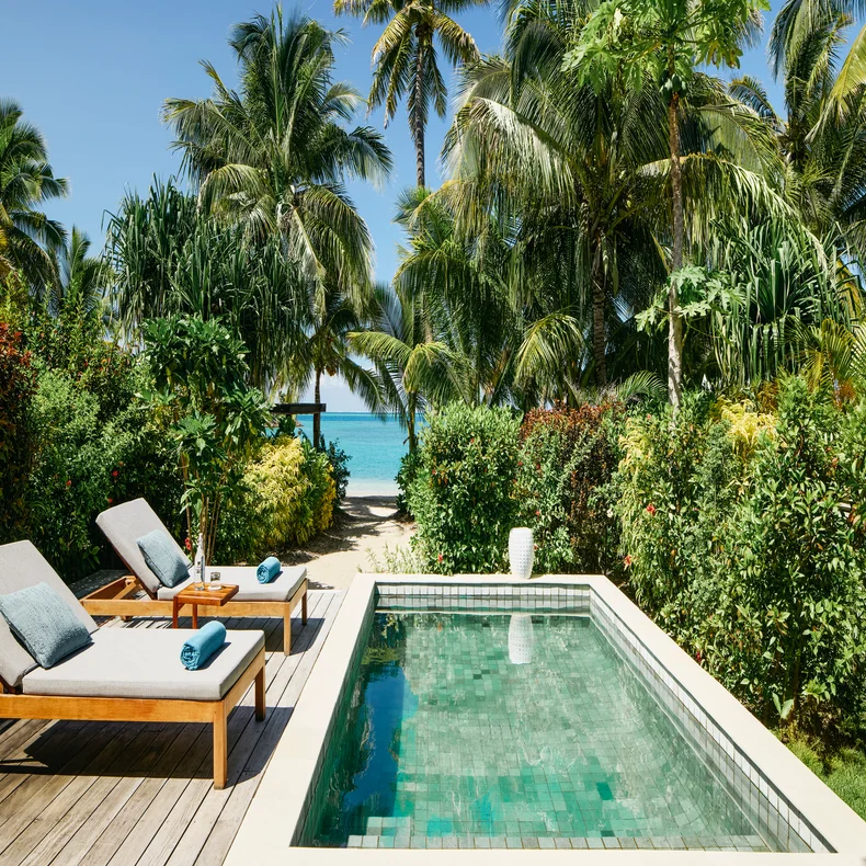 Private pool bordered by tropical greenery, wooden deck with lounge chair, overlooking beach, palm trees, and clear blue ocean beyond.