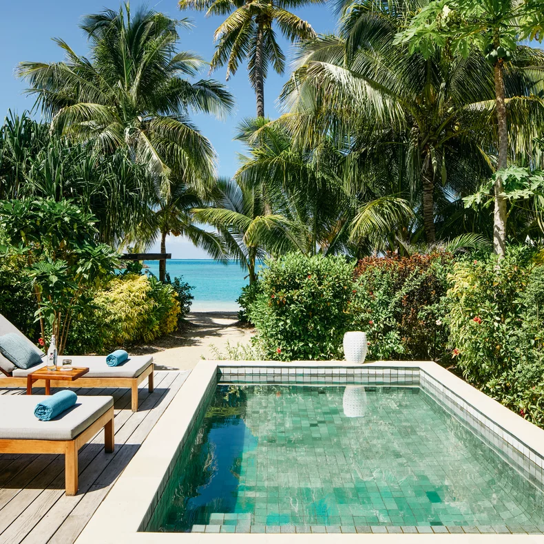 Private pool bordered by tropical greenery, wooden deck with lounge chair, overlooking beach, palm trees, and clear blue ocean beyond.