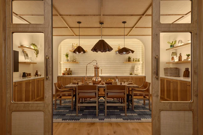 Dining area with natural wood table and chairs, pendant lighting, open shelving with greenery and ceramics, and a geometric rug beneath the table.
