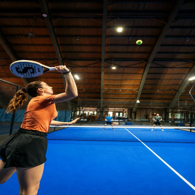 Un client en tenue de sport joue au padel sur un court couvert bleu, dans un cadre soigneusement conçu pour favoriser le bien-être et un mode de vie actif.