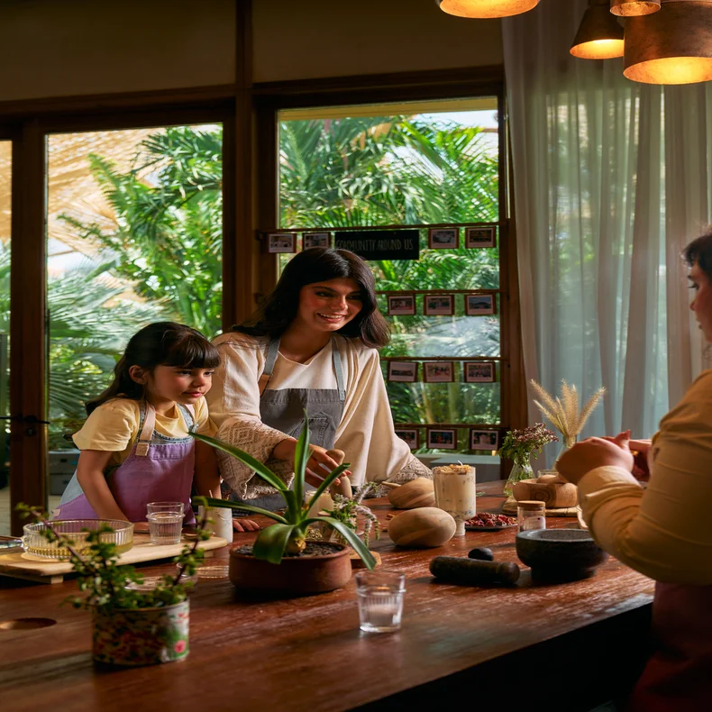 Dos personas con delantales preparan juntas una comida en una mesa de madera cubierta de macetas de barro y plantas verdes. Esta luminosa estancia está bañada por la luz natural del sol, y la ventana da a un paisaje verde.
