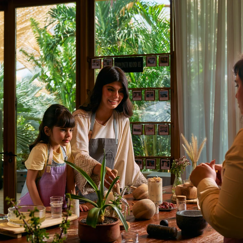 Dos personas con delantales preparan juntas una comida en una mesa de madera cubierta de macetas de barro y plantas verdes. Esta luminosa estancia está bañada por la luz natural del sol, y la ventana da a un paisaje verde.