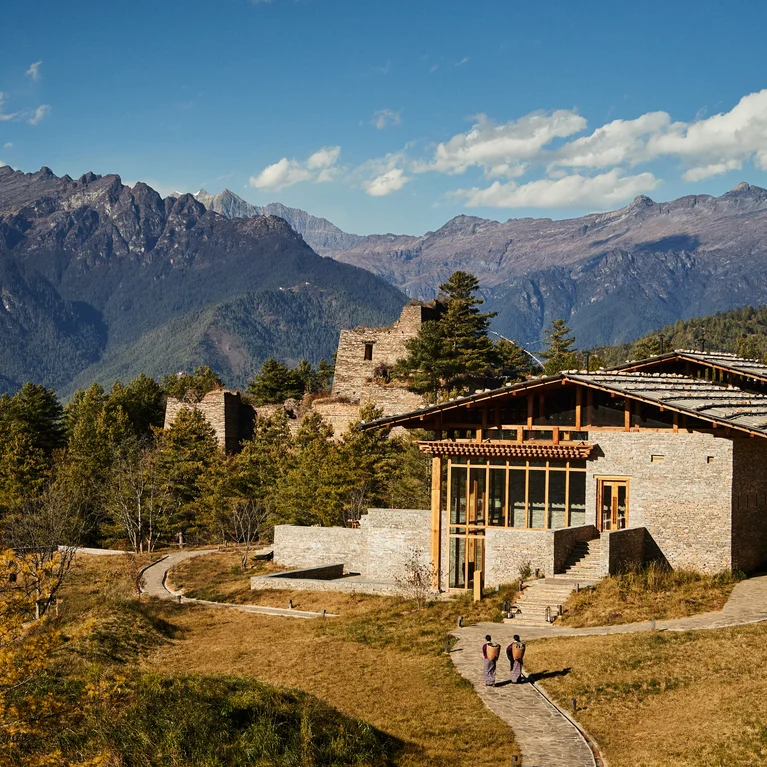 A stone building surrounded by trees and hills, with two people walking along a path; forested mountains rise in the distance beneath a blue sky.