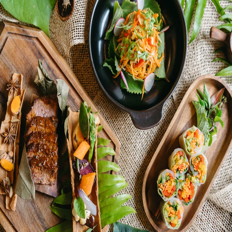 Grilled meat, spring rolls, and a vibrant salad presented on wooden plates with fresh green leaves and herbs, highlighting local ingredients and mindful presentation.