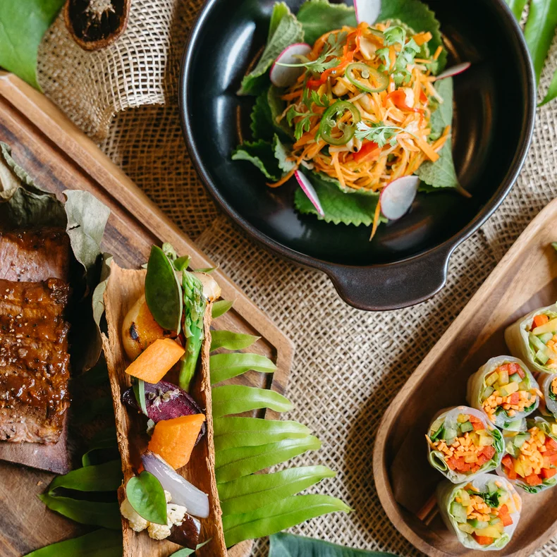 Grilled meat, spring rolls, and a vibrant salad presented on wooden plates with fresh green leaves and herbs, highlighting local ingredients and mindful presentation.
