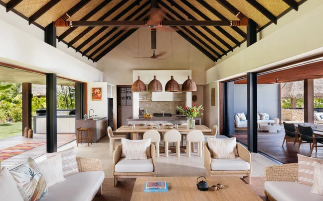Spacious living area featuring two beige sofas, dark wood coffee table, large windows providing natural light, and views of palm trees and ocean coastline.